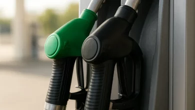 The image features a close-up of a fuel pump at a gas station, focusing on two nozzles, one with a green handle and another with a black handle. The nozzles are positioned prominently on the right side of the frame, while the background remains blurred, hinting at a bright, well-lit environment typical of a gas station. The lighting is soft and natural, enhancing the colors of the nozzles, which contrast with the metallic surface of the pump. The overall mood is functional and commercial, cap…
