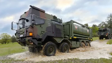 На зображенні зображено військовий вантажівка, що проїжджає через невелику затоплену ділянку дороги.