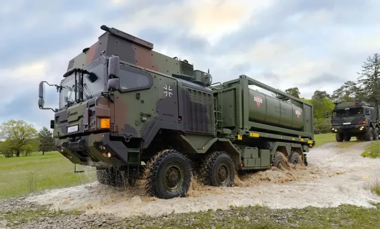 На зображенні зображено військовий вантажівка, що проїжджає через невелику затоплену ділянку дороги.