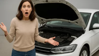 A surprised woman stands in front of a car with its hood open, positioned slightly angled towards the camera.